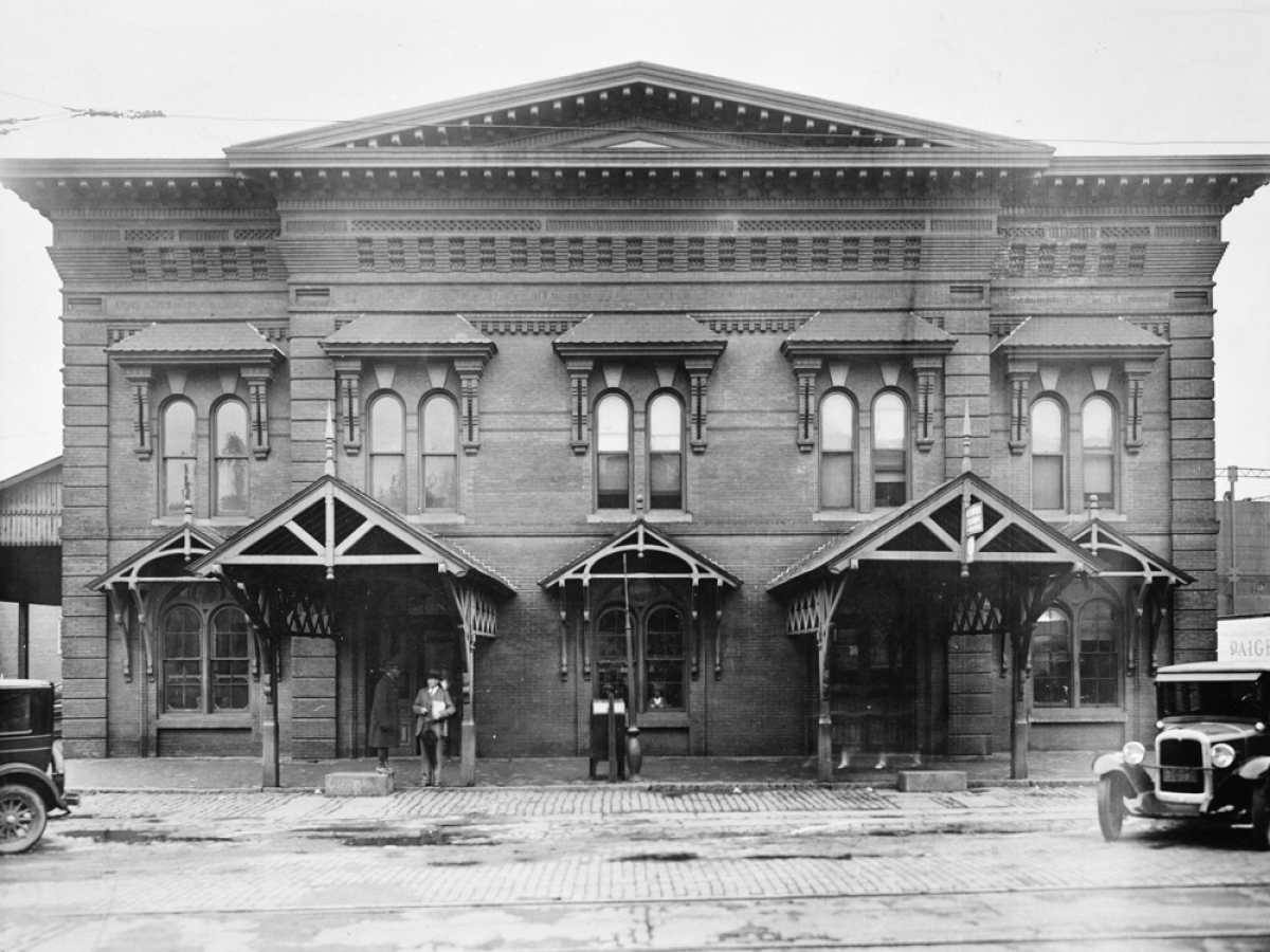 Historic brick train station with three archways and vintage cars in front.