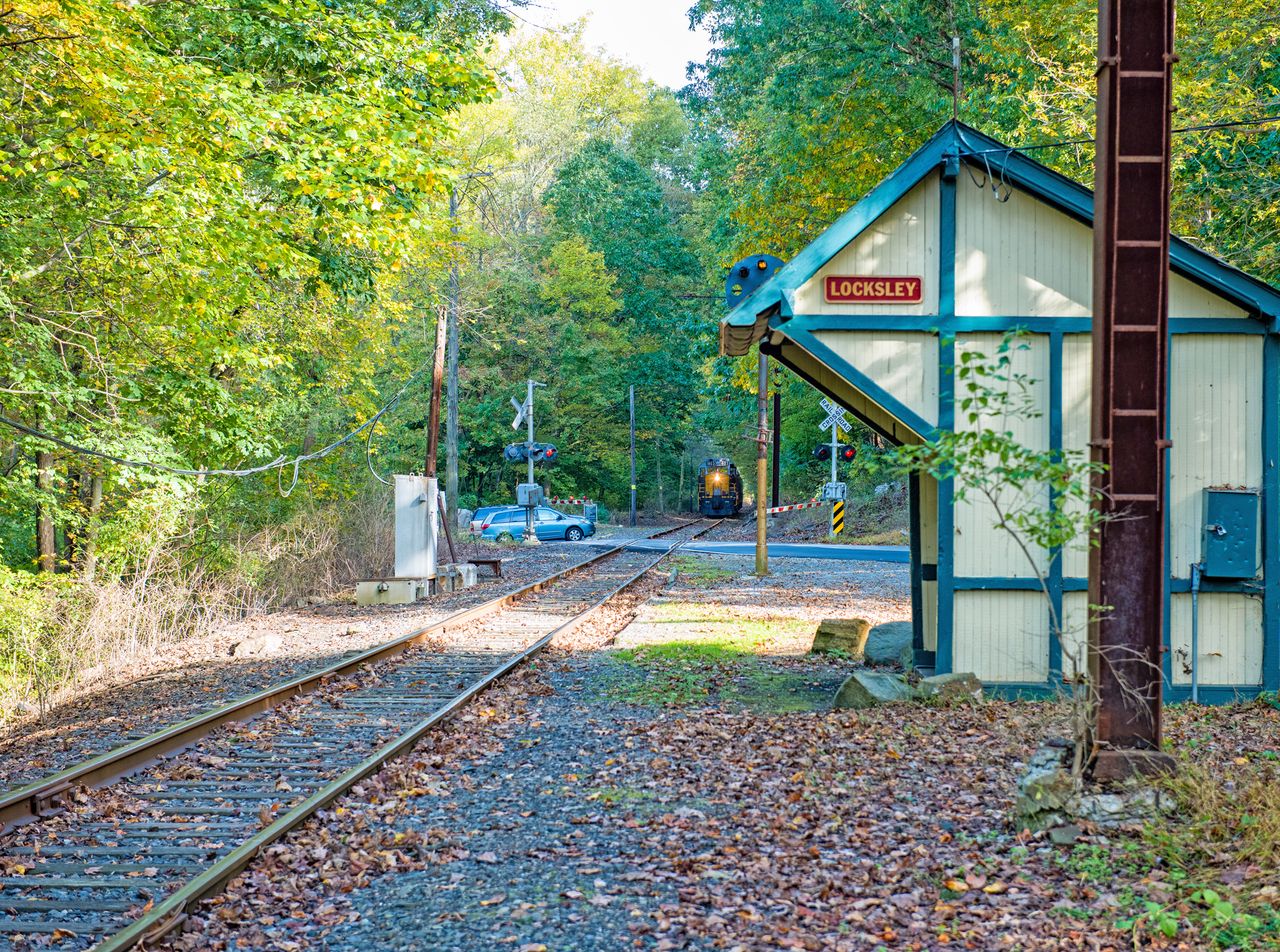 Small train station with tracks, trees, and a distant train in the background.