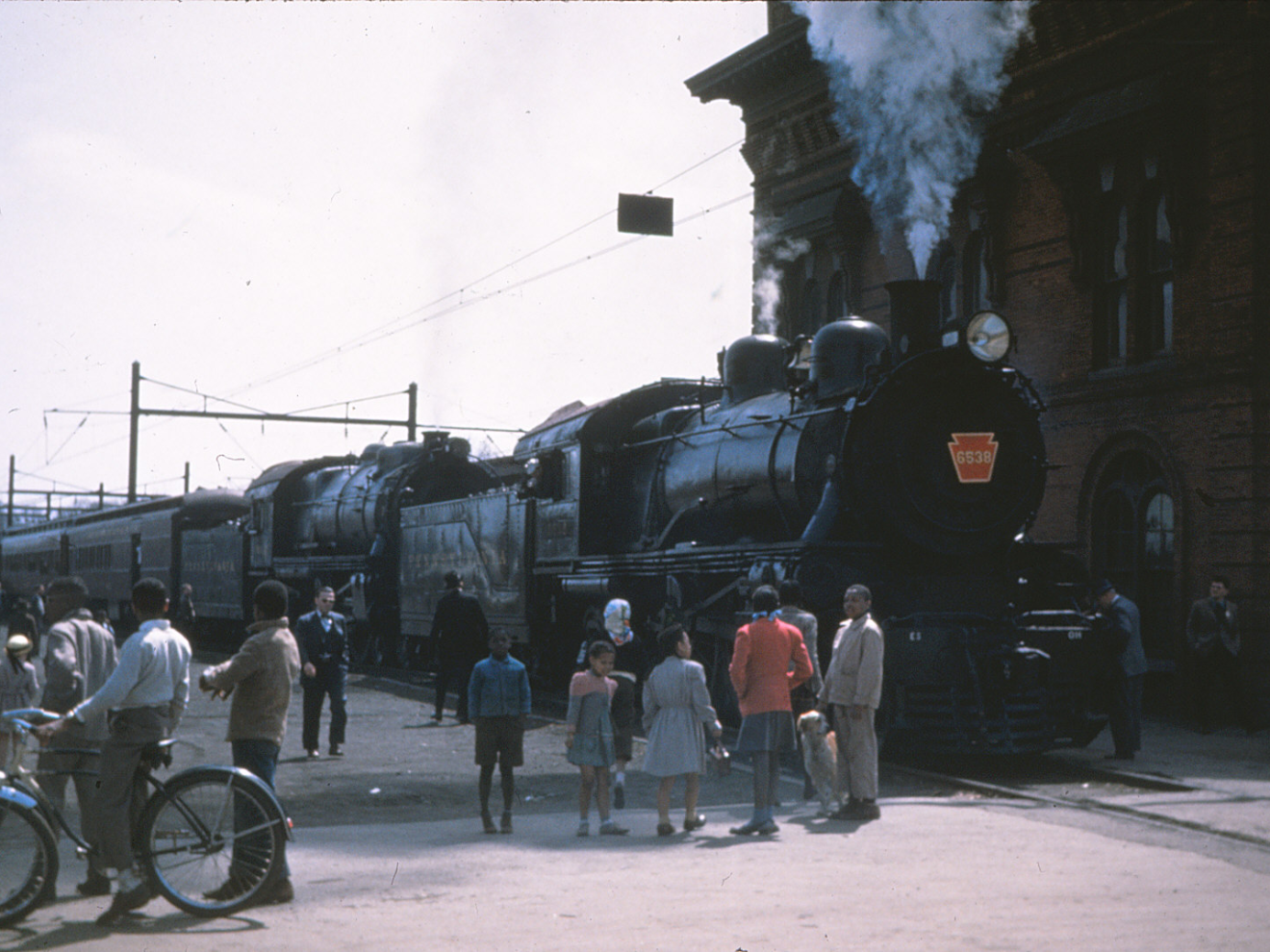 Steam locomotives at a station with people and bicycles nearby.